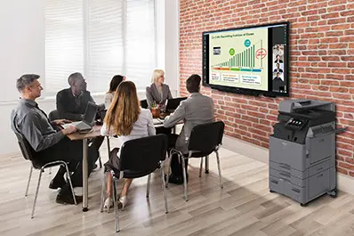 Meeting in a conference room with people viewing a video conference and presentation on a collaboration display and a copier machine in the foreground.